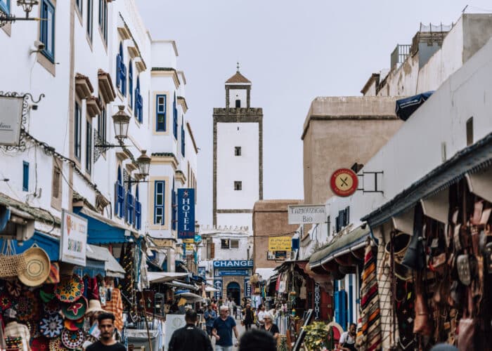 Busy market street in Essaouira medina with white buildings and mosque tower in Morocco