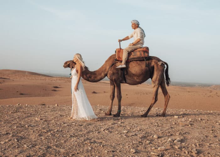 15 Day Morocco Tour from Marrakech to Casablanca – Man riding camel and woman in white dress standing in desert landscape during golden hour